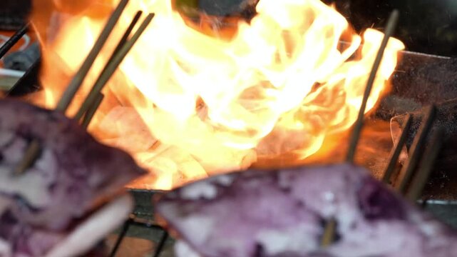 Close-up 4K footage of fresh squid grilled on hot iron plate with big orange flame fire, flaming seafood barbecue street food cooking with smoke.
