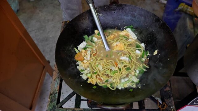 Close-up 4K footage of Indonesian Javanese noodle soup mie godog mie rebus cooking and stir frying in black wok with cabbage carrot egg and broth, traditional Asian street food.