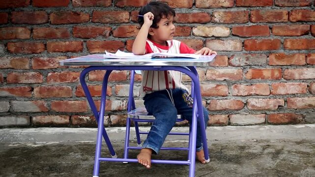 Elementary age cute child of Indian ethnicity sitting on chair near brick wall at home and studying book and writing alphabets in notebook video portrait close up.