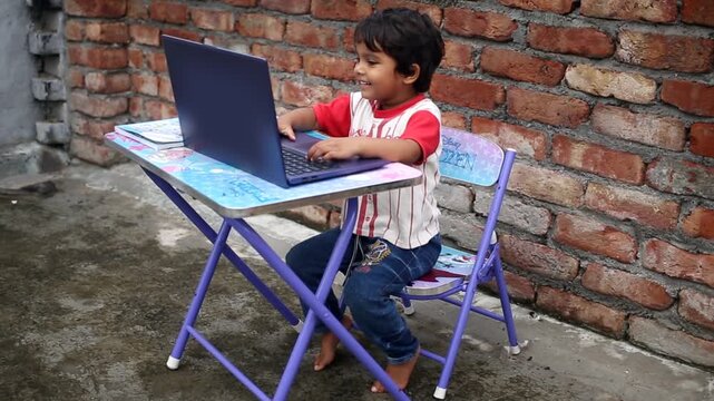 Elementary age cute child of Indian ethnicity sitting on chair near brick wall at home and studying book and writing alphabets in notebook video portrait close up.