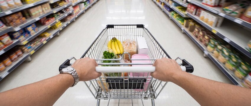 A first-person perspective shot of hands pushing a shopping cart filled with groceries down a supermarket aisle.
