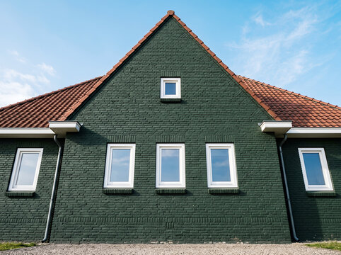 Exterior view of a dark green brick house with a gabled roof, red clay tiles, and white-framed windows against a clear blue sky