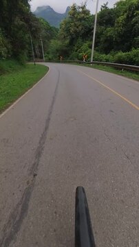 Vertical cyclist POV riding on a winding mountain road through lush forest, with guardrails, chevron signs, utility poles and distant mountains in the background.