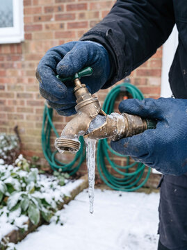 Gloved hands holding a burst frozen brass garden tap with dripping icicle