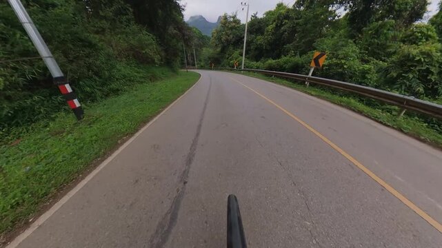Cyclist POV riding along a winding mountain road through lush green forest, with guardrails, utility poles and scenic mountain views under an overcast sky.
