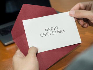 Person receives Merry Christmas greeting card with red envelope near laptop at a work desk. Holiday wishes, seasonal correspondence, festive communication and Christmas greetings concepts. © tete_escape