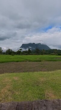 Vertical timelapse of dramatic mountains surrounded by clouds above green rice fields and rural farmland, capturing changing weather and peaceful tropical scenery.
