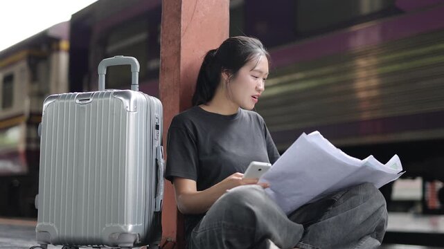 Young Asian woman sitting on the platform floor at a railway station. Traveler planning her journey with a map and a smartphone.