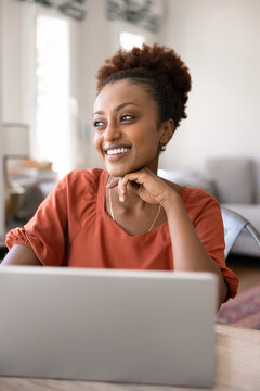 Young African woman sitting at desk with laptop, gazing to side with smile, exuding satisfaction with completion of remote study or workflow. Modern technology usage for leisure, business or learning