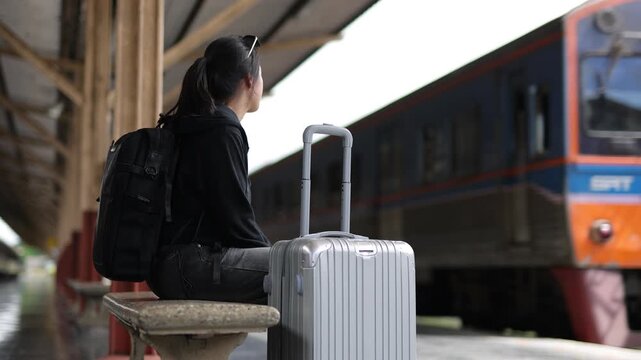 Young Asian traveler sitting on a bench with her backpack and suitcase, patiently waiting on the platform as a train passes by.