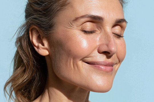 Mature woman close up face with closed eyes smiling in soft sunlight against pastel blue background, natural skin texture and joyful calm expression