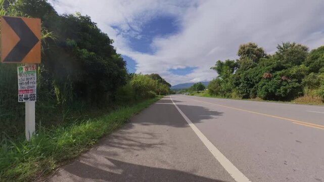 Scenic rural road surrounded by lush green vegetation, with distant mountains and dramatic clouds under a partly blue sky, creating a peaceful countryside landscape.