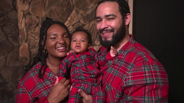 Smiling young parents hold their baby son in matching red plaid Christmas pajamas. Happy African American family portrait by a stone fireplace wall. Camera slowly pushes in on joyful faces.