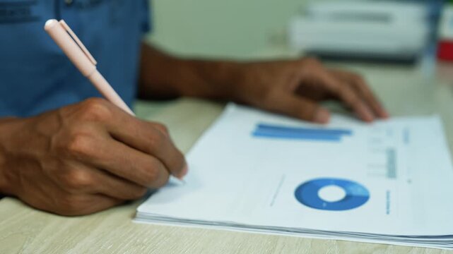 Healthcare worker writing medical report with charts, Close-up of a healthcare worker in blue scrubs writing on a medical report with charts and a blue pie graph at a desk.