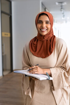 Young adult Indian woman wear hijab and abaya holding planner, writing notes, organizing work tasks in modern office, posing for professional corporate portrait. Business administration, management