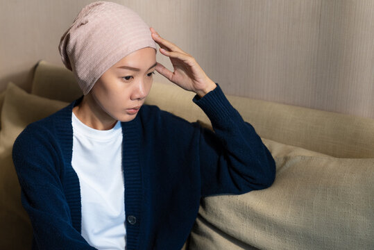 Thoughtful Asian woman wearing a headscarf touching her temple while sitting on the floor. Cancer patient, mental stress, chemotherapy recovery, and health concept.