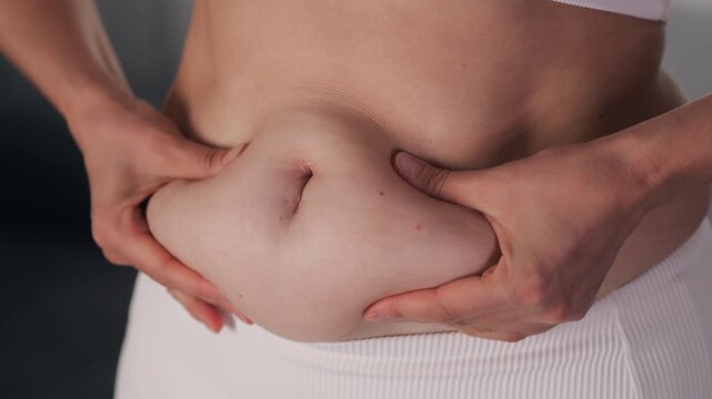 Close-up of woman pinching excess fat and skin around her abdomen and waist, illustrating body fat, weight management and body changes.