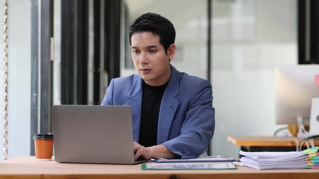 Professional businessman working intently at desk, reviewing documents and taking notes while using laptop, concentrated effort in corporate.