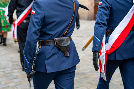 Close-up rear view of dark blue police dress uniform with Polish flag patch on sleeve, black leather pistol holster on belt, and white and red sash during ceremonial parade.   