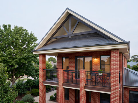 Red brick house exterior with an upper balcony, dark metal roof, black railings, and a gabled roofline with timber beams, residential architecture