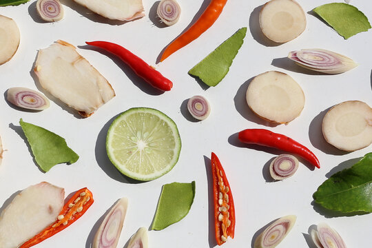 A colorful, close-up pattern of Thai herbs: lime slices, galangal, lemongrass, chilies, and kaffir lime leaves.