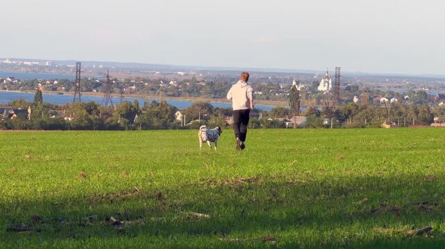 Young man running with open arms together with spotted dalmatian dog on green field outdoors. Active pet owner expressing joy and freedom in countryside landscape near lake on sunny day.