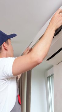 Young Male Technician Inspects Broken Home Appliance During Indoor Maintenance Work. Focused Serviceman Checks Faulty Air Conditioner Unit Carefully, Troubleshooting Ventilation System Indoors.