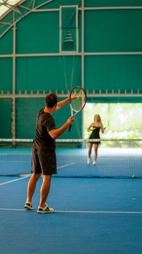 A man preparing to serve a tennis ball on an indoor blue court with a partner in the background