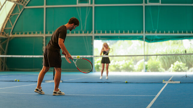 A man preparing to serve a tennis ball on an indoor blue court with a woman waiting in background