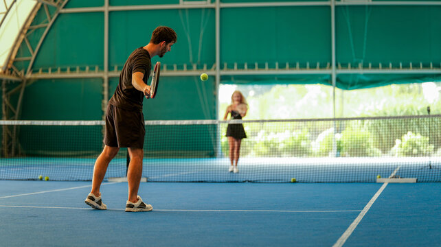 A man playing tennis on an indoor blue court with a woman waiting on the opposite side of the net