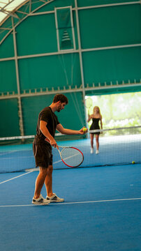 Young man holding a tennis ball and racket on an indoor blue court with a partner in the distance