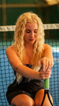 Young blonde woman with wavy hair resting on a tennis racket at an indoor court facility