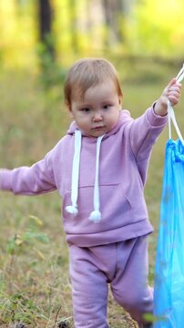 Little Child Carries Large Blue Trash Bag While Walking Through Autumn Forest. Dedicated Toddler Helps With Outdoor Cleanup, Showing Curiosity And Helpfulness Among Green Trees And Fallen Leaves.