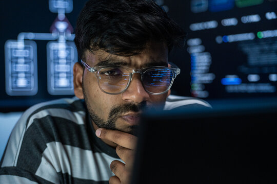 Indian software developer stares intently at computer screens while writing code in a dark room. The focused programmer feels determination and concentration while solving complex digital problems.