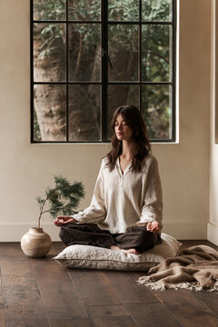 A woman meditates on a linen cushion in a cozy contemporary bedroom. Mindfulness, stress reduction and self-care concept