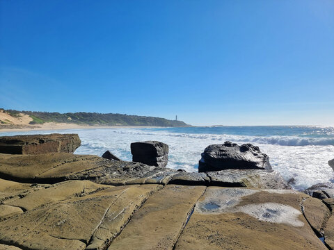 Weathered rock shelves and sculpted boulders at Soldiers Point at low tide, with breaking surf and distant Norah Head Lighthouse on the Central Coast of New South Wales, Australia.
