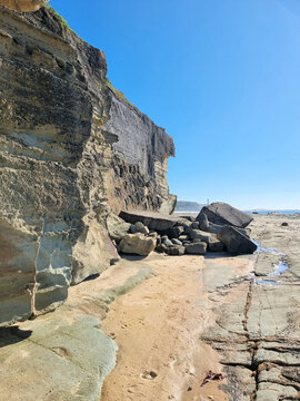 Fallen boulders beneath a weathered, layered coastal cliff at Soldiers Point, with sand and shallow pools exposed at low tide on the Central Coast of New South Wales, Australia.