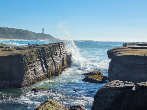 Waves crash through a channel between weathered rock platforms at Soldiers Point, with Norah Head Lighthouse in the distance on the Central Coast of New South Wales, Australia.