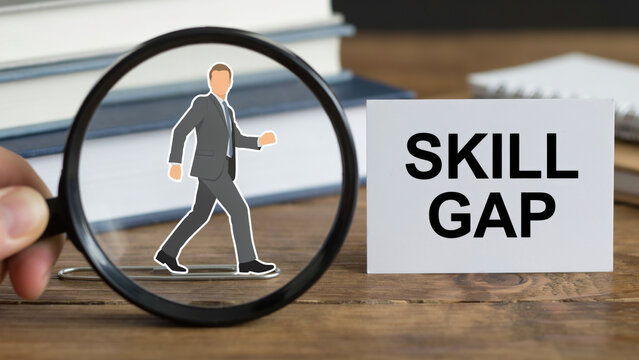 A hand holding a magnifying glass over a paper businessman figure next to a card with the text skill gap on a wooden desk with cutout businessman placed uses magnifying