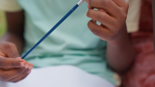 Child hands manipulate blue paintbrush near white drawing surface. Background shows blurred figures and objects indicating group setting. Soft natural light illuminates scene emphasizing