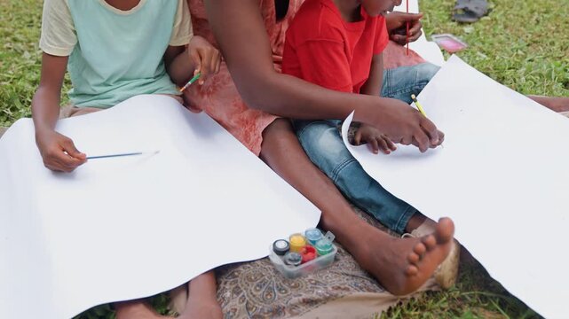 Children and adult participate in painting activity using brushes and pencils on large white paper with paint containers visible. They sit outdoors on patterned fabric surrounded by grass