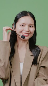 A friendly Asian woman wearing a headset speaks to a customer against a green background. Great for call center, customer service, and online support videos.