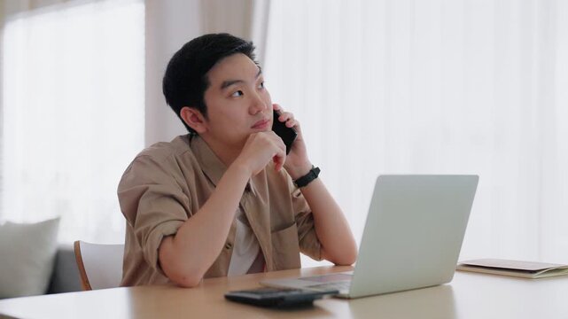 Happy young Asian man working on laptop in living room. Freelance worker male sitting at desk writing notes while watching webinar, studying online