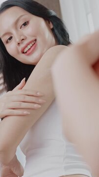An Asian woman looking in a mirror and gently applying body lotion to her arm.