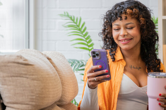 cheerful young woman with curly hair using smartphone, smiling with satisfaction while relaxing at home, texting on mobile phone