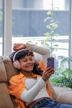 Young African American woman on the sofa at home, listening to music with a smartphone and headphones or speaking on a live stream
