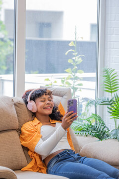 Young African American woman on the sofa at home, listening to music with a smartphone and headphones or speaking on a live stream