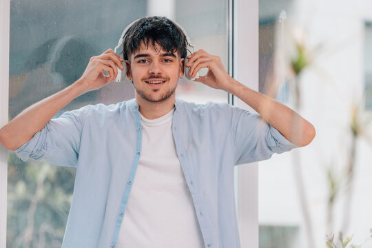 Young man smiling while putting on white wireless headphones near a window, wearing casual clothes, enjoying music in a bright urban setting