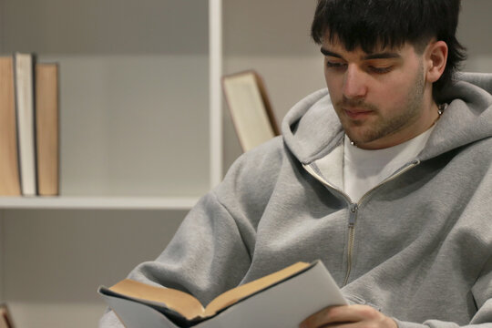 young man in a gray hoodie reads a book while sitting near a bookshelf indoors, in a calm and cozy setting