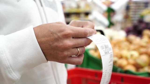 Hands hold receipt near produce stall. Person wears white jacket, has wedding ring. Background shows onions and shopping basket. Scene suggests retail shopping or grocery checkout